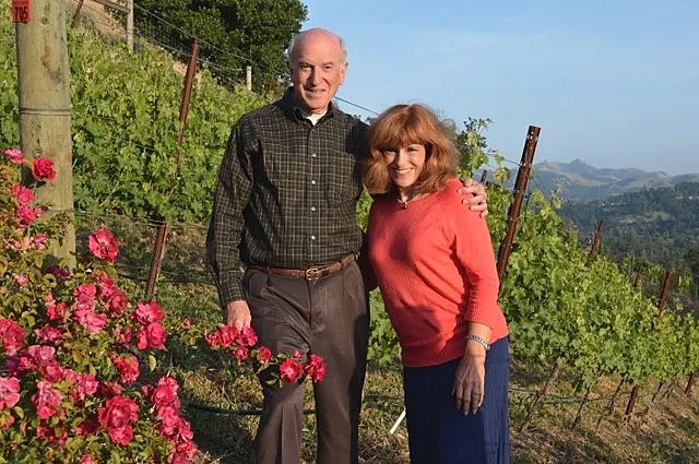 Larry and Esther Thal standing together at the edge of the vineyard, with vine rows and hills behind them.